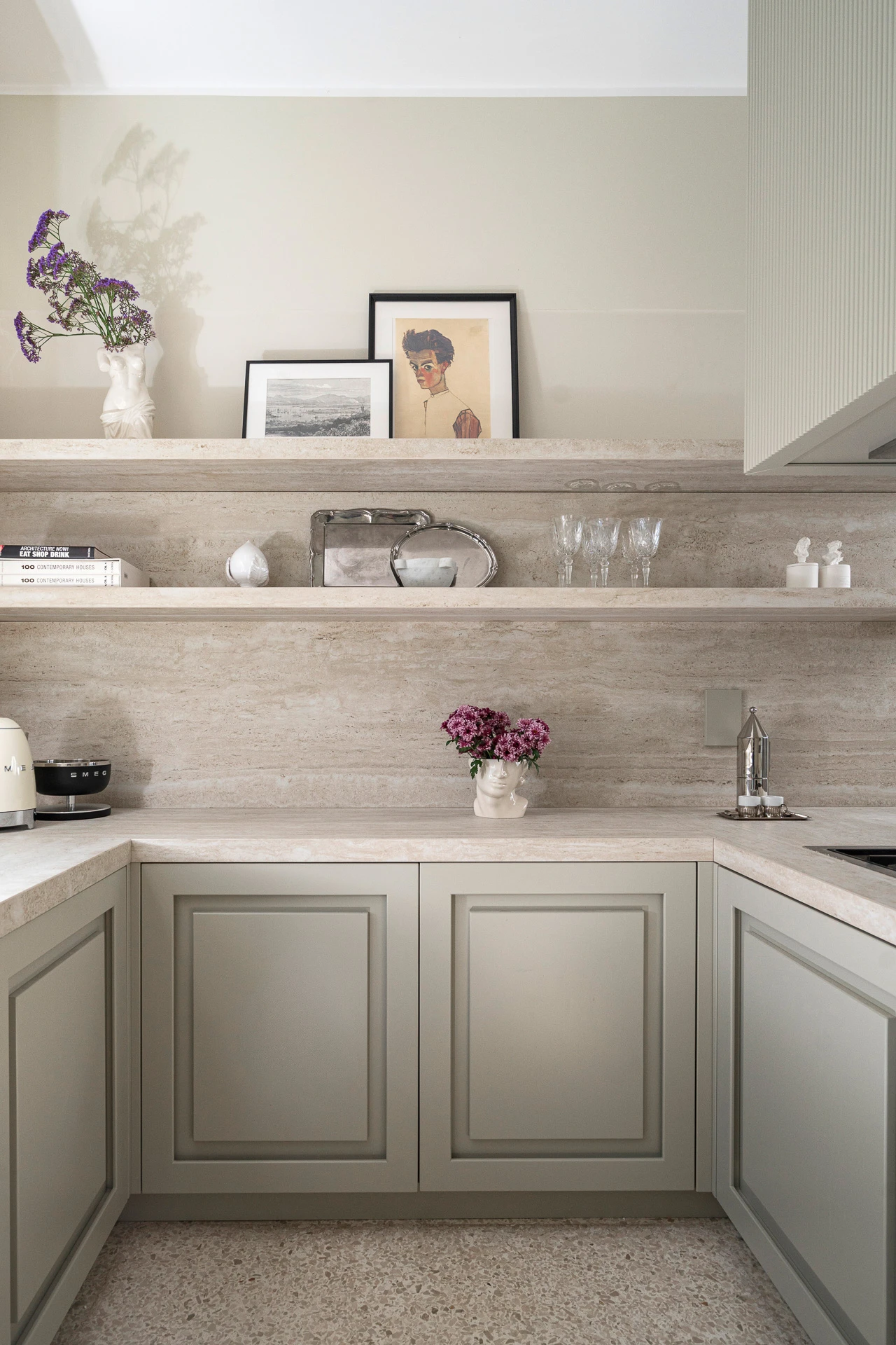 Custom corner kitchen with sage green framed doors, travertine countertop and cladding, and decorated open shelves
