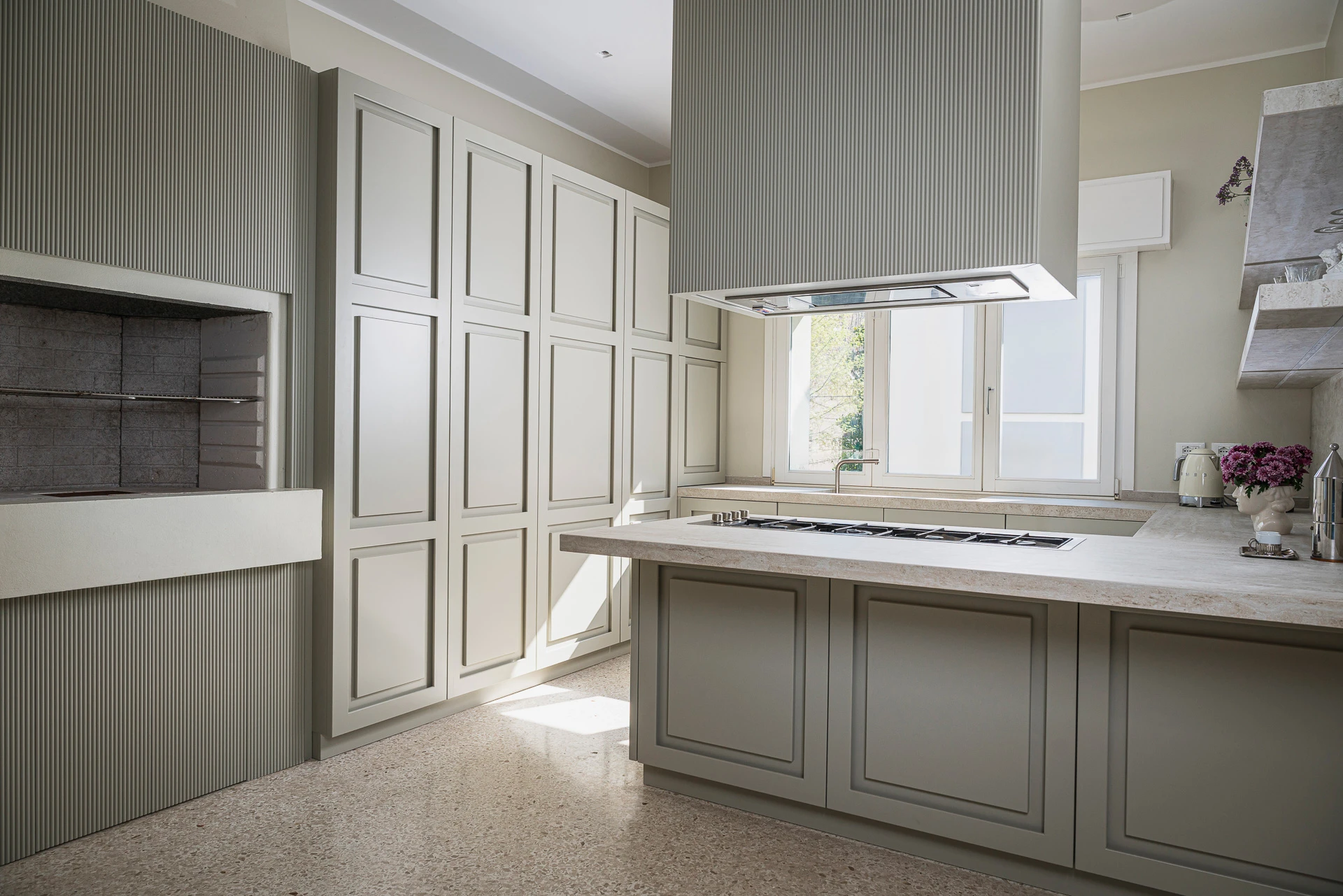 Progettazione di cucina su misura: isola con piano cottura, dispensa a colonne e camino a legna integrato nell'ambiente